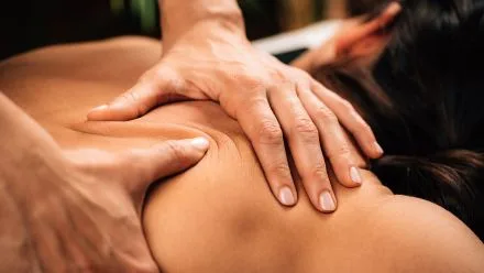Therapist's hands applying deep massage pressure to a person's upper back and shoulder while they lie face down.