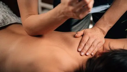 Therapist's hands pressing the upper back and shoulder of a person lying face down on a massage table.