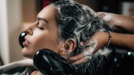 Woman reclining with eyes closed as a stylist massages shampoo foam into her hair over a salon sink.