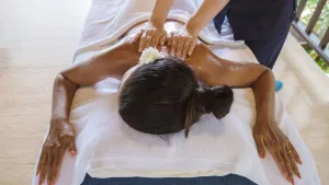 Woman lying face down on a massage table receiving an oiled back and shoulder massage, hair in a bun with a white flower.