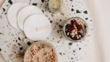 Top-down view of cosmetic jars and round cotton pads on terrazzo surface, with loose powder and dried flower petals nearby.