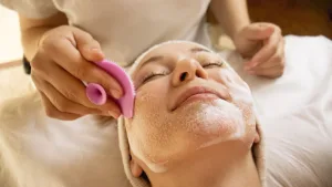 Woman lying with eyes closed while a cosmetician scrubs foamy facial cleanser using a pink silicone brush.