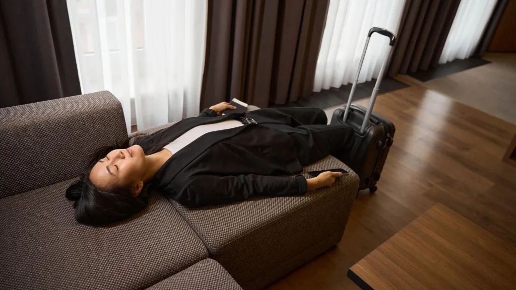 Woman in business attire lying back on a hotel sofa with suitcase and phone nearby, eyes closed and appearing rested.