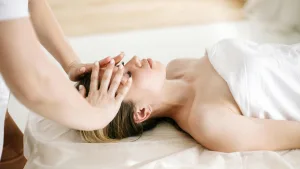 Woman lying on a massage table with a white sheet, receiving a gentle forehead and temple massage from a therapist's hands.
